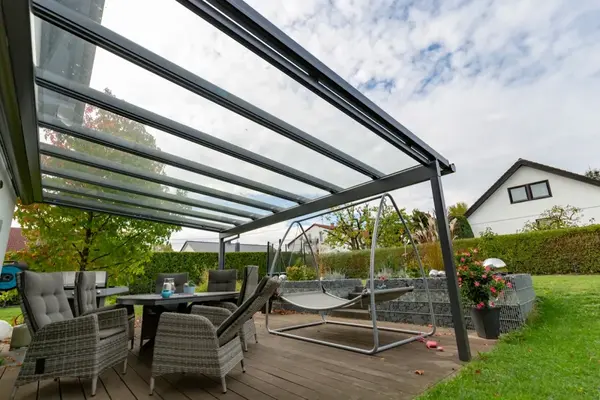 Moderne Terrassenüberdachung aus Glas mit Gartenmöbeln und Hängeliege im gepflegten Garten.