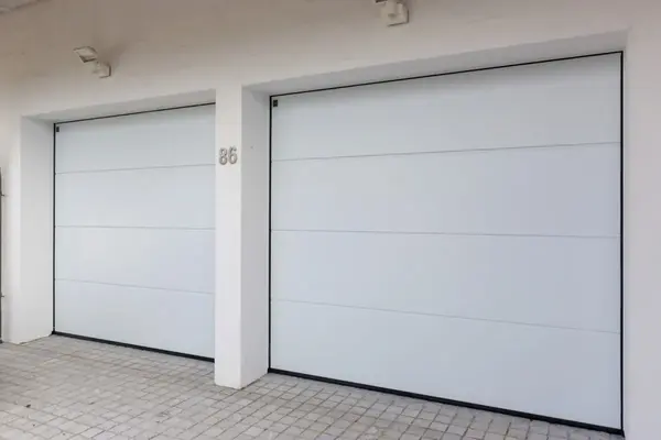 Zwei moderne, weiße Garagentore in einer Hausfassade mit Pflastersteinen im Vordergrund.